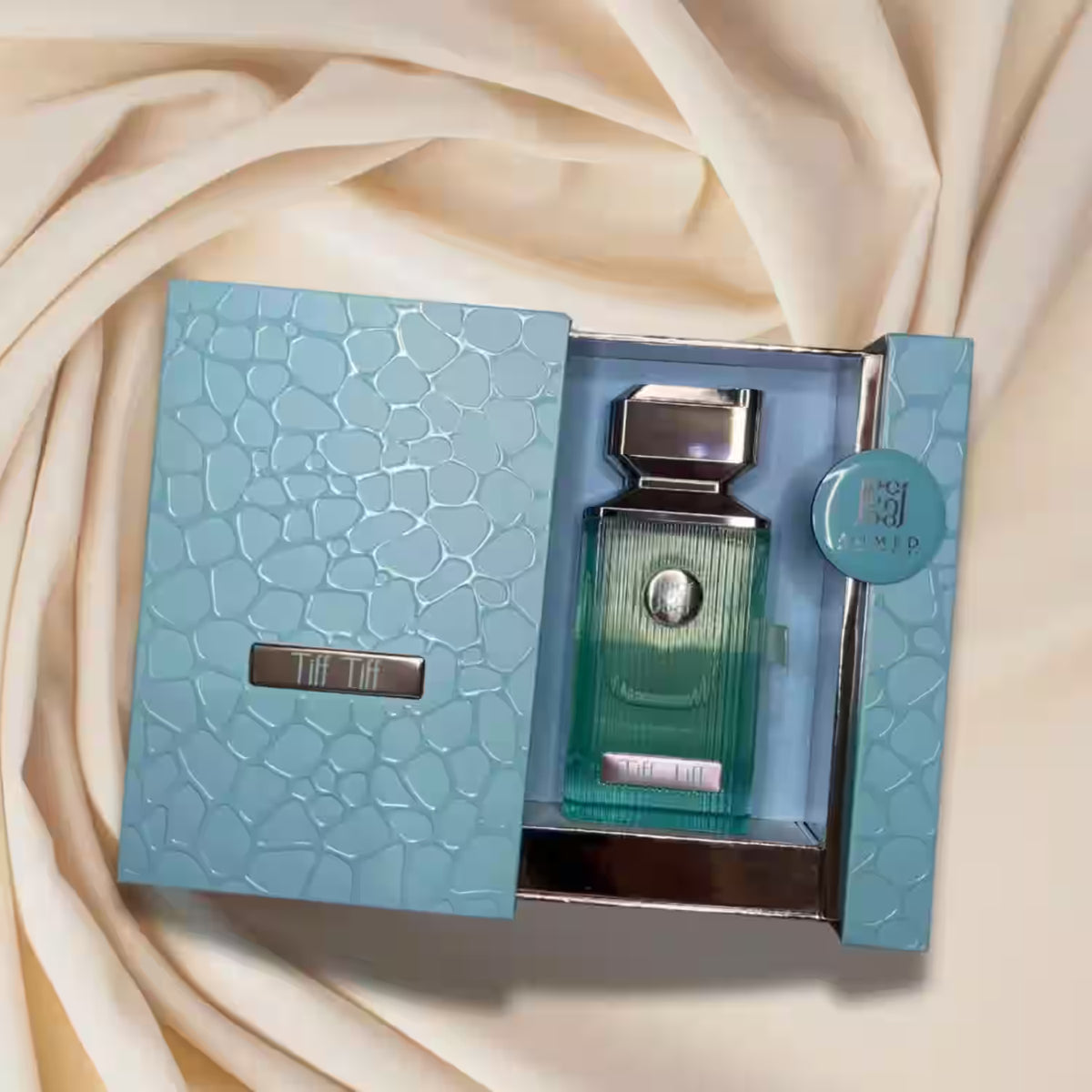 A perfume bottle with a ribbed translucent green-blue body and a silver cap, presented in an open textured light blue box on draped cream fabric.