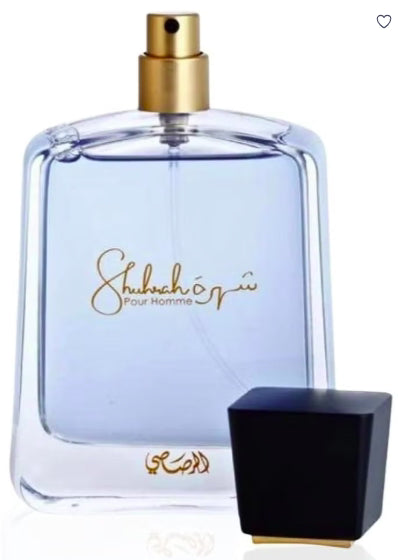 A clear glass perfume bottle with pale blue liquid and a gold spray top, shown alongside a black cap with a small gold base. The bottle has gold script text reading 'Shuhbah Pour Homme' on a white background.